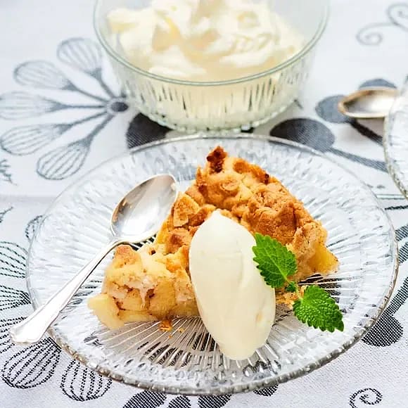 En bit äppelpaj med vaniljglass och mynta på en glasskiva, med vispad grädde i bakgrunden.