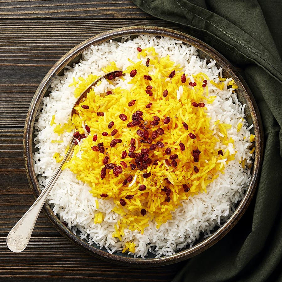 Kryddad ris med saffran och torkade bär serverat i en skål, omgiven av vitt ris och en silverbestick.