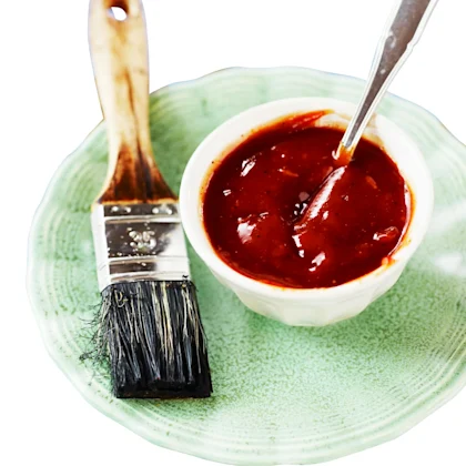 BBQ sås i en vit skål som står på en grön tallrik, en sked i såsen. Bredvid skålen ligger en pensel.