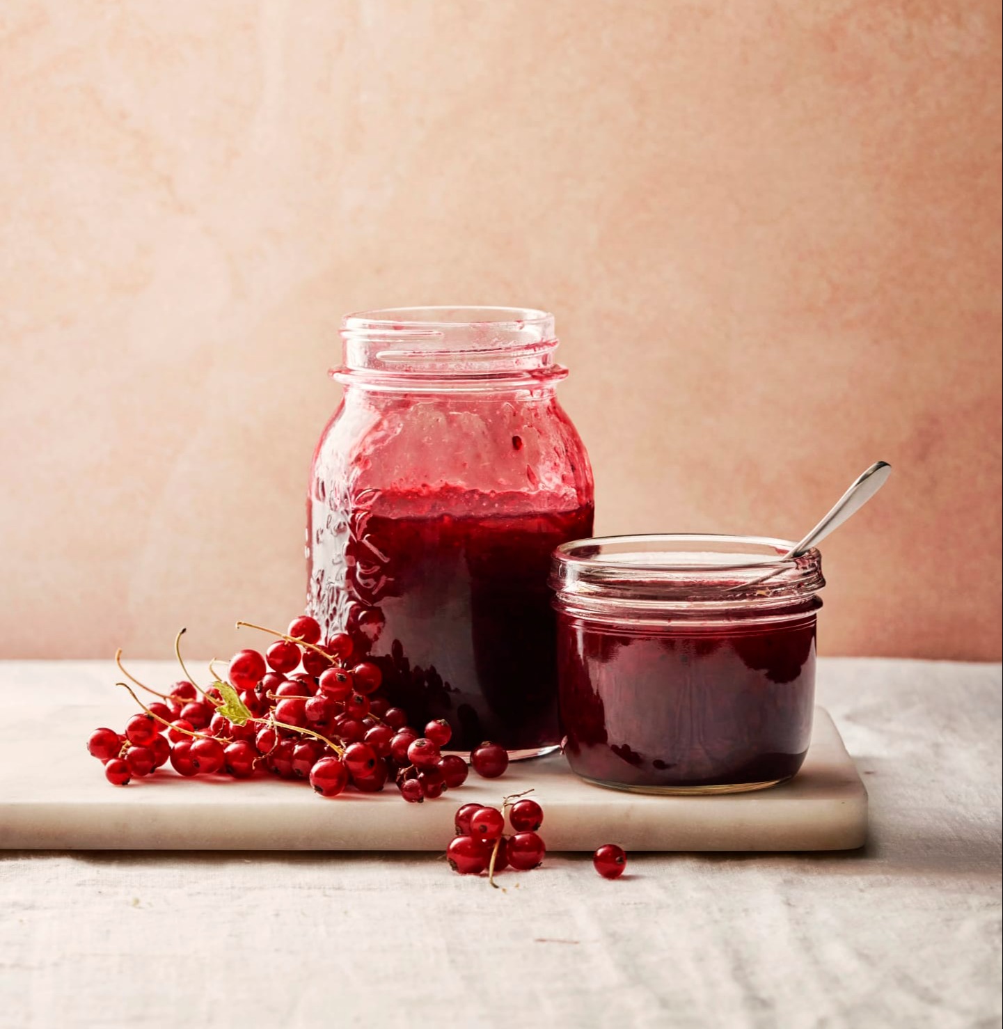 Två glasburkar med vinbärssylt står på en skärbräda, omgivna av färska vinbär. Bakgrunden är mjukt rosa.