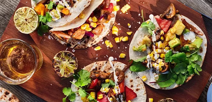 Tacos med grillade grönsaker, majs, avokado, koriander och dressing på tortillabröd, serverade på en skärbräda med grillad lime och dryck bredvid.