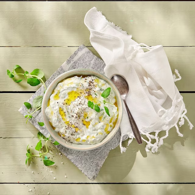 Skål med tzatziki toppad med svartpeppar, olivolja och färska örter, placerad på en stenplatta med en sked och en vit servett bredvid.