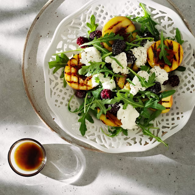 Sallad med grillade persikor, björnbär, ruccola och klickar av färskost på ett fat, bredvid ett litet glas med dressing.