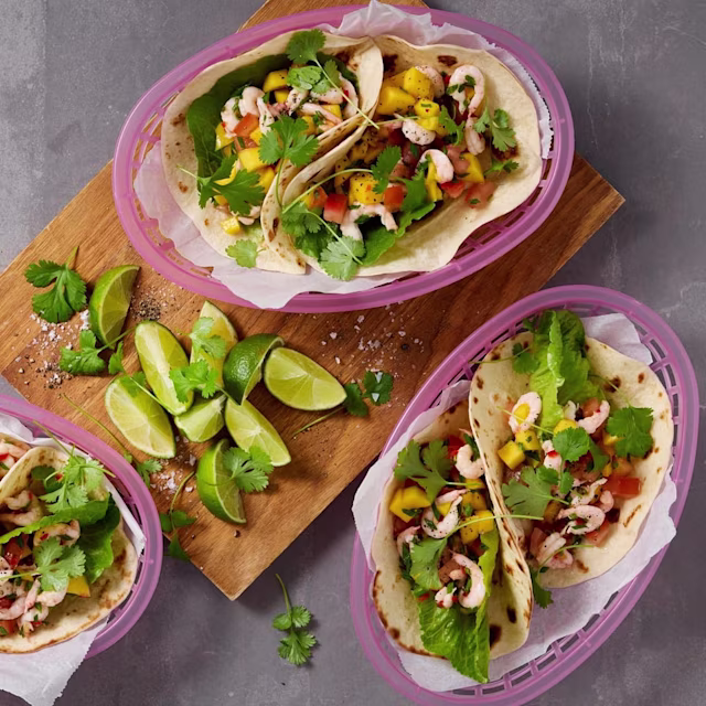 Tacos med räkor, mango, sallad, koriander och grönsaker i mjuka tortillabröd, serverade i rosa korgar med limeklyftor och koriander på en träbricka.