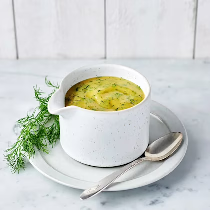 Krämig sås med dill i vit kanna på tallrik med sked och dillkvist på marmorbord.