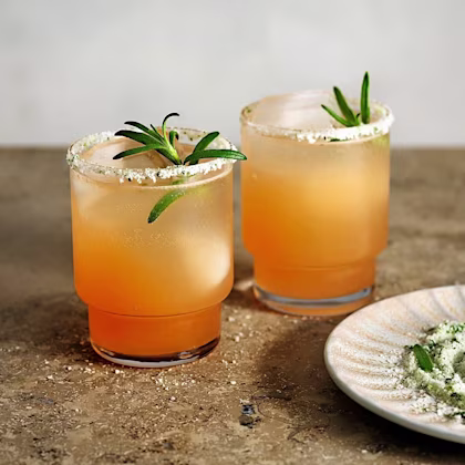 Två orangea drinkar med is och rosmarinkvistar, glasens kanter är doppade i salt och örter.