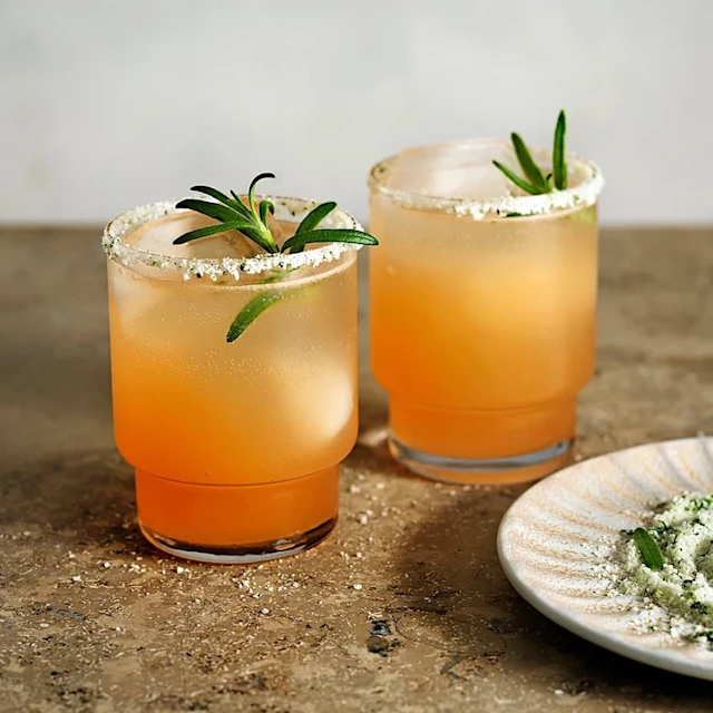 Två orangea drinkar med is och rosmarinkvistar, glasens kanter är doppade i salt och örter.