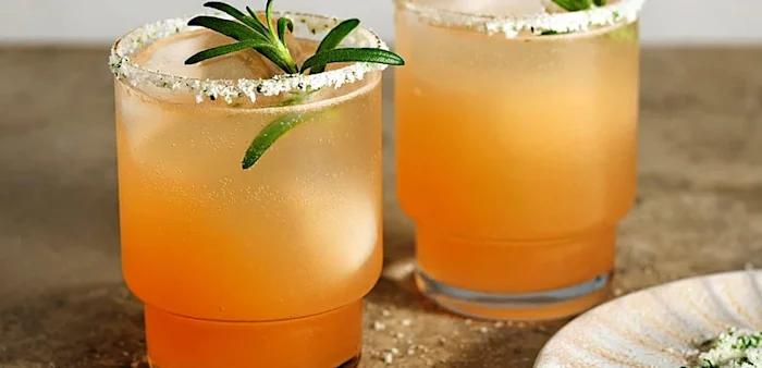 Rosa cocktail i cocktailglas med äppelskiva, jordgubbar och rhubarberstjälkar på bord med shaker i bakgrunden.