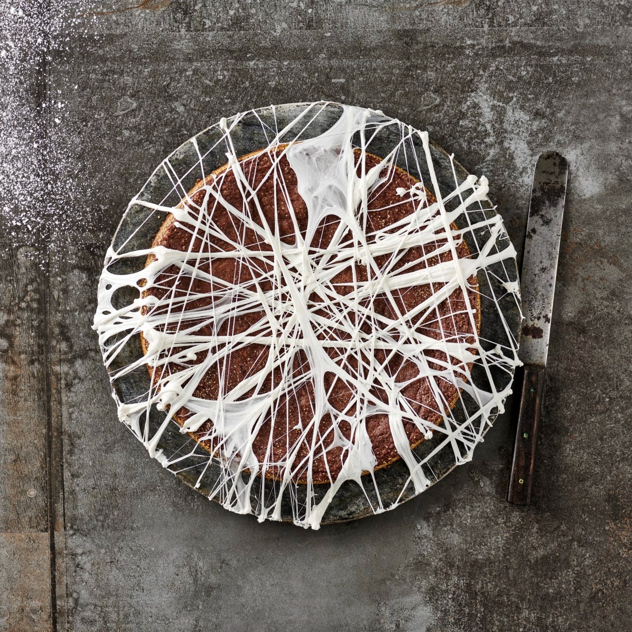  Kladdkaka dekorerad med vita trådar av smält marshmallow på ett fat, med en kniv bredvid och florsocker på bordet