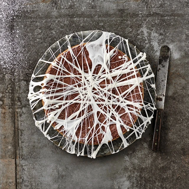 Kladdkaka dekorerad med vita trådar av smält marshmallow på ett fat, med en kniv bredvid och florsocker på bordet