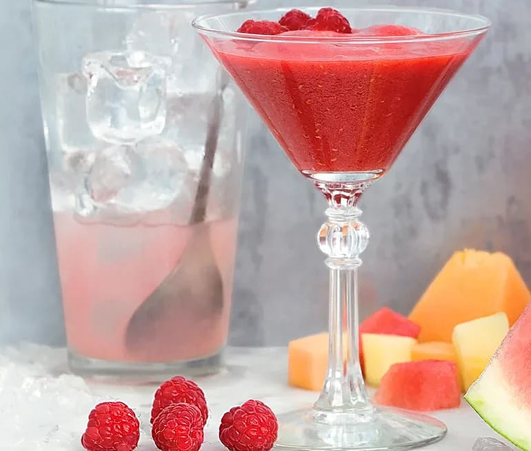 Röd bärdrink i cocktailglas med hallon, isbitar i glas med sked och skuren melon och vattenmelon i bakgrunden.