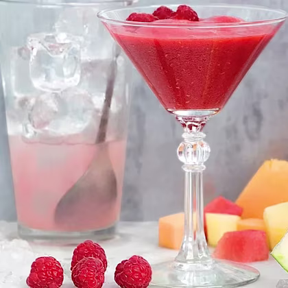 Röd bärdrink i cocktailglas med hallon, isbitar i glas med sked och skuren melon och vattenmelon i bakgrunden.
