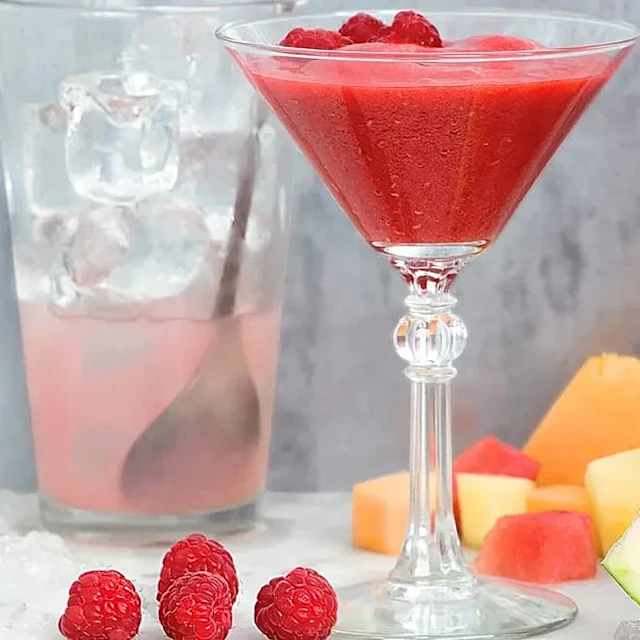 Röd bärdrink i cocktailglas med hallon, isbitar i glas med sked och skuren melon och vattenmelon i bakgrunden.