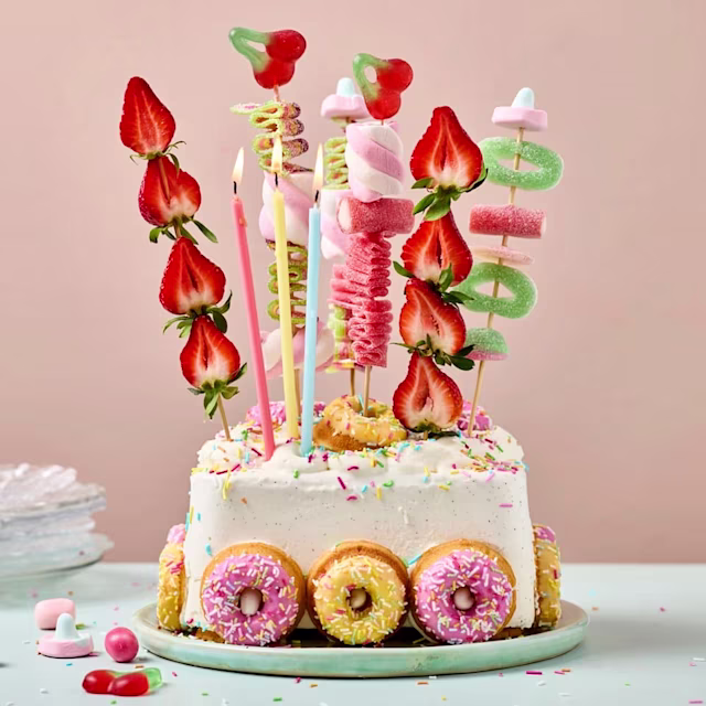 Fyrkantig tårta dekorerad med små donuts runt kanten, färgglada strössel, jordgubbar, godis och marshmallows på pinnar samt tre tända ljus på toppen.