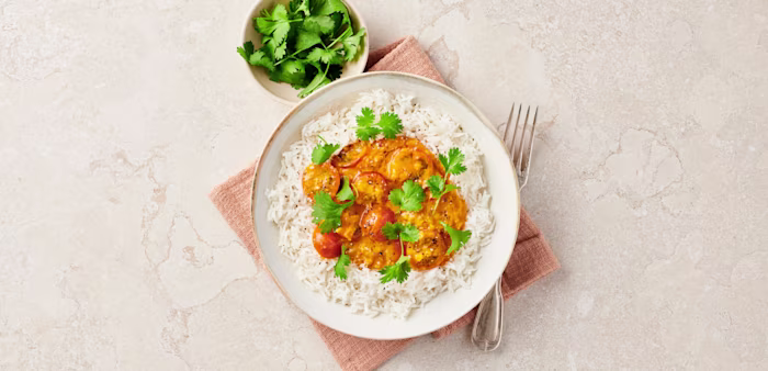 Ris med gryta av räkor i tomat- och currysås, toppat med koriander, serverat med en skål färsk koriander och gaffel.