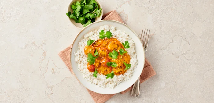 Ris med gryta av räkor i tomat- och currysås, toppat med koriander, serverat med en skål färsk koriander och gaffel.