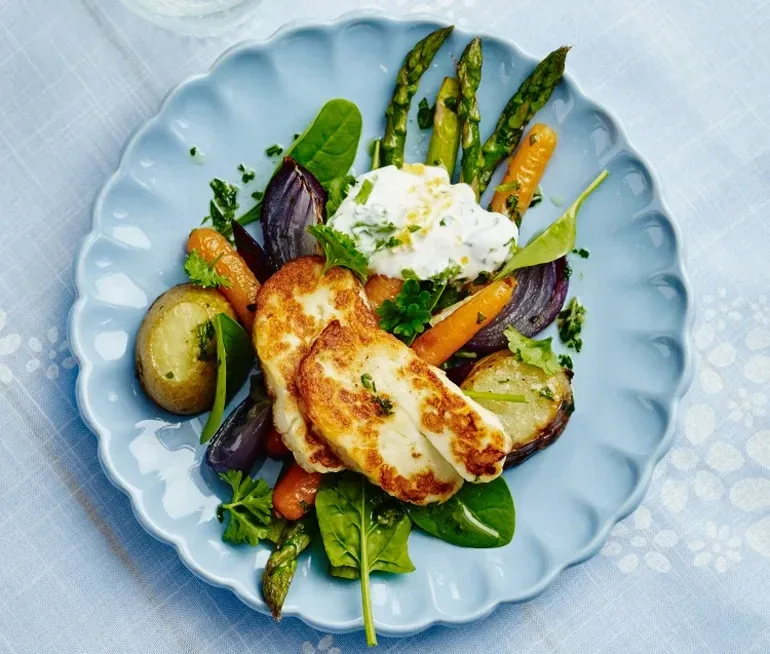 Grillad halloumi med ugnsrostade grönsaker som sparris, morötter, rödlök, potatis och spenatblad, toppat med en klick yoghurtsås på ett blått fat.