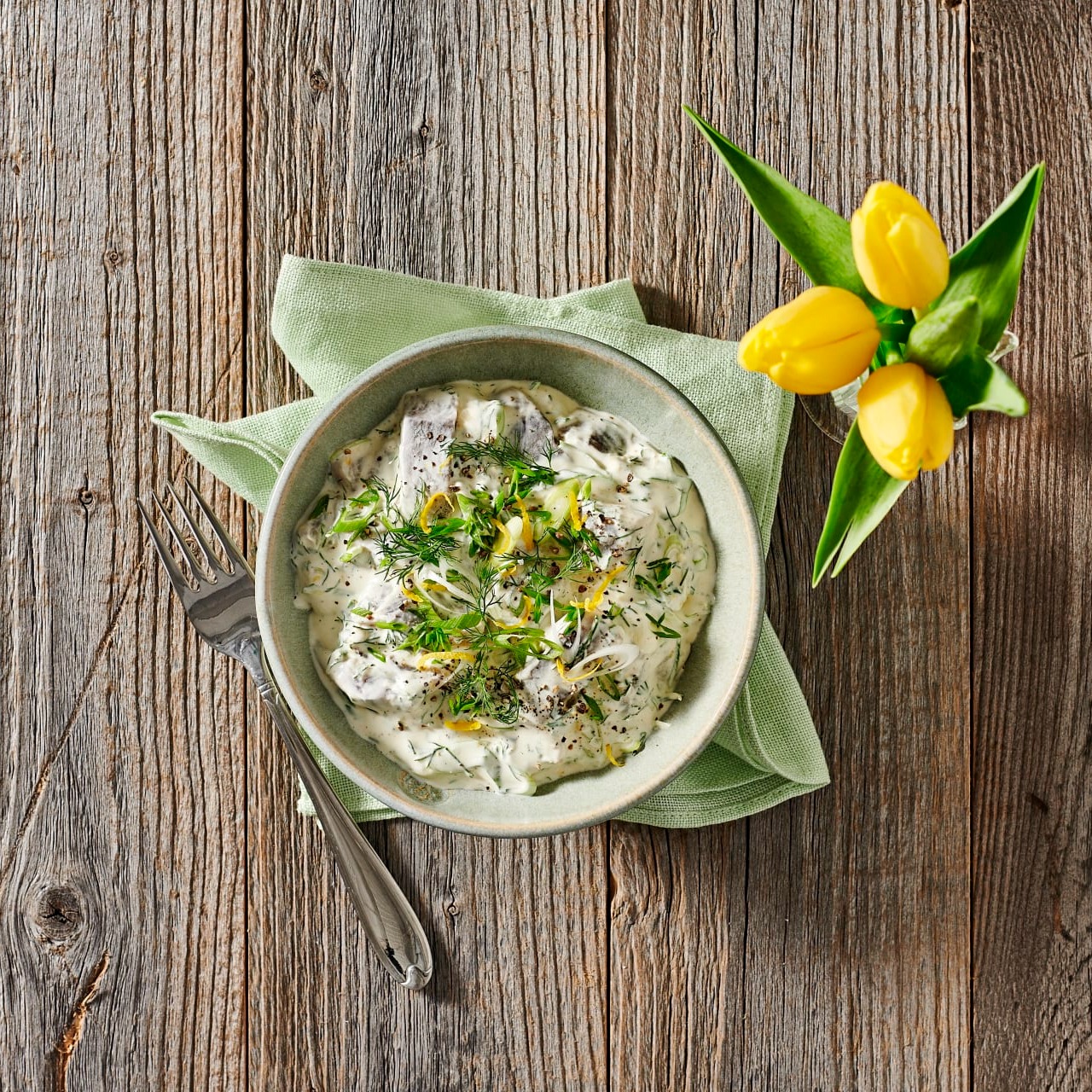 Påsksill i krämig sås med dill och citron i en skål, serverad med en gaffel och grön servett, samt en vas med gula tulpaner.