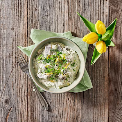 Påsksill i krämig sås med dill och citron i en skål, serverad med en gaffel och grön servett, samt en vas med gula tulpaner.