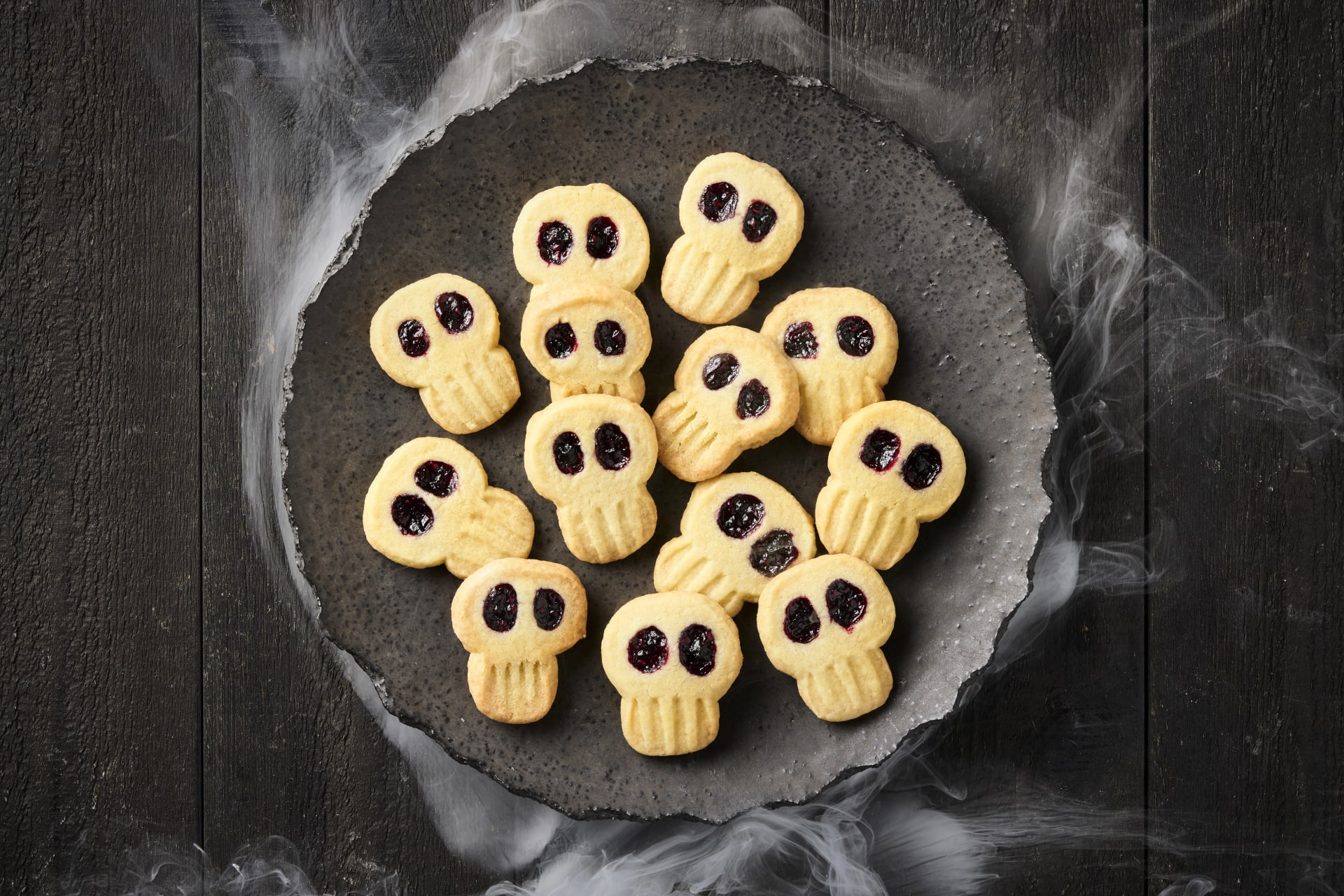 Småkakor formade som döskallar med mörka ögon av sylt, upplagda på ett fat med rök runt