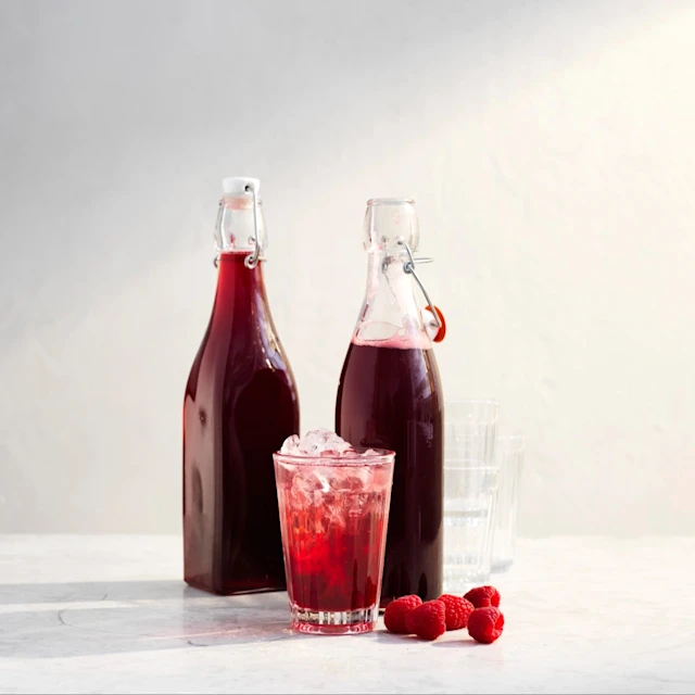 Hallonsaft i ett glas med is, omgiven av två flaskor hallonsaft och färska hallon på bordet.