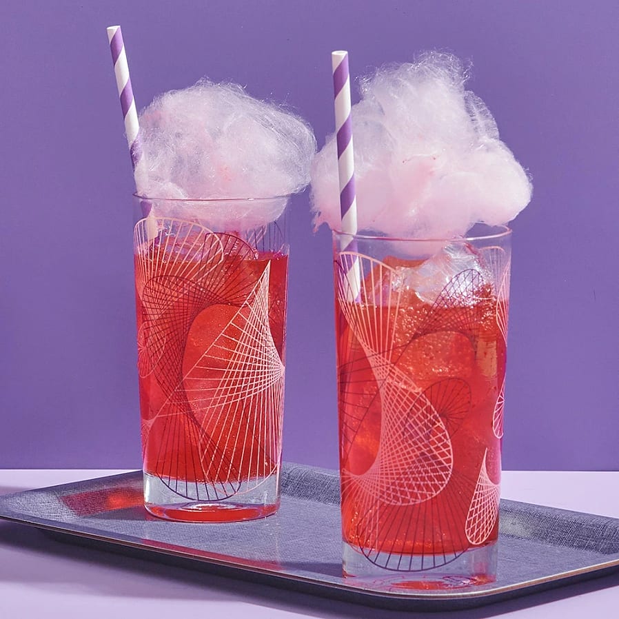 Två glas med röd dryck och is, toppade med rosa sockervadd och randiga sugrör på bricka.