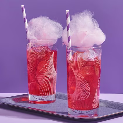 Två glas med röd dryck och is, toppade med rosa sockervadd och randiga sugrör på bricka.