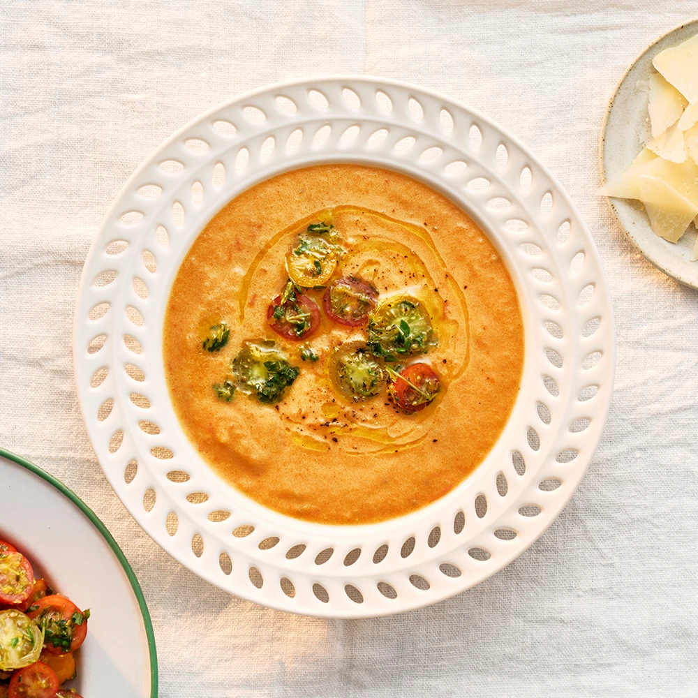 Soppa toppad med halverade tomater, örter och ringlad olja i en djup tallrik på en duk.