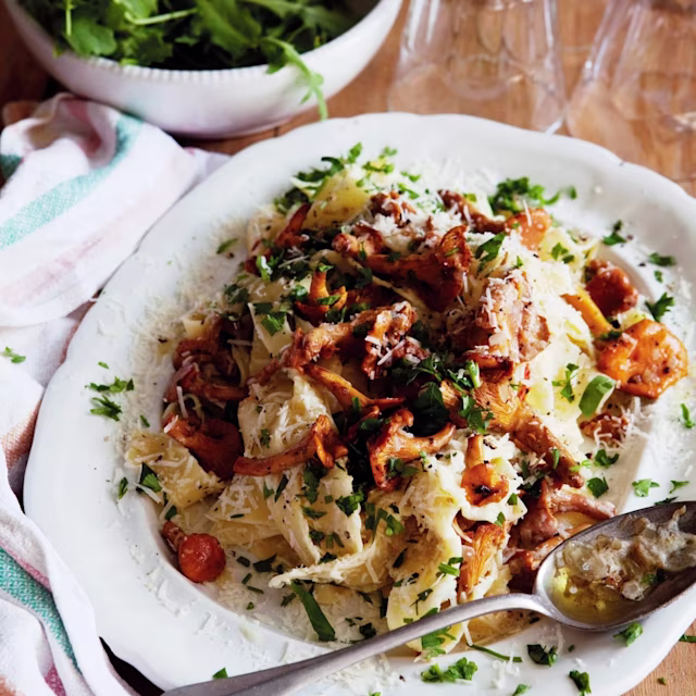 Pasta med kantareller, parmesanost och färsk persilja, serverad på en vit tallrik. Grön sallad syns i bakgrunden.