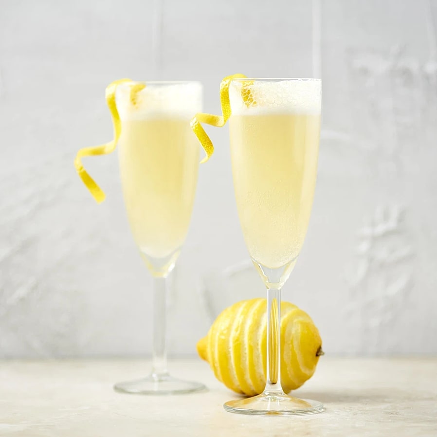 Två champagneglas med skummande citroncocktail och citronskal som dekoration, bredvid en hel citron.