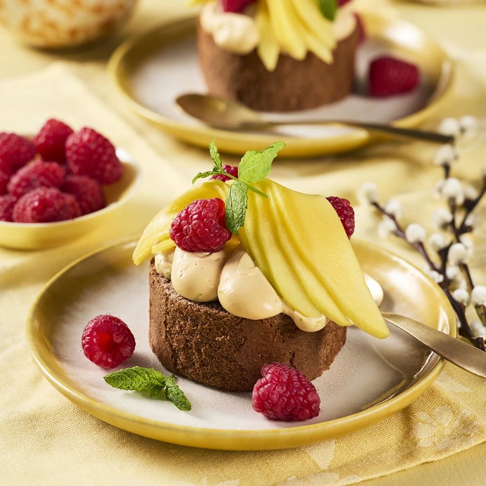 Chokladbakelse toppad med kräm, skivor av mango, hallon och mynta på en assiett, med fler hallon i en skål bredvid.