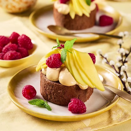 Chokladbakelse toppad med kräm, skivor av mango, hallon och mynta på en assiett, med fler hallon i en skål bredvid.