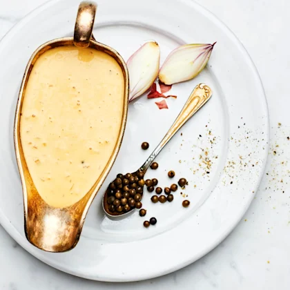 Grönpepparås i en skål som står på en vit tallrik. På tallriken ligger 2 stycken scarlottenlökar samt en sked med grönpeppar i. Det ligger 4 stycken scharlottenlökar med skal på till vänster om tallriken.