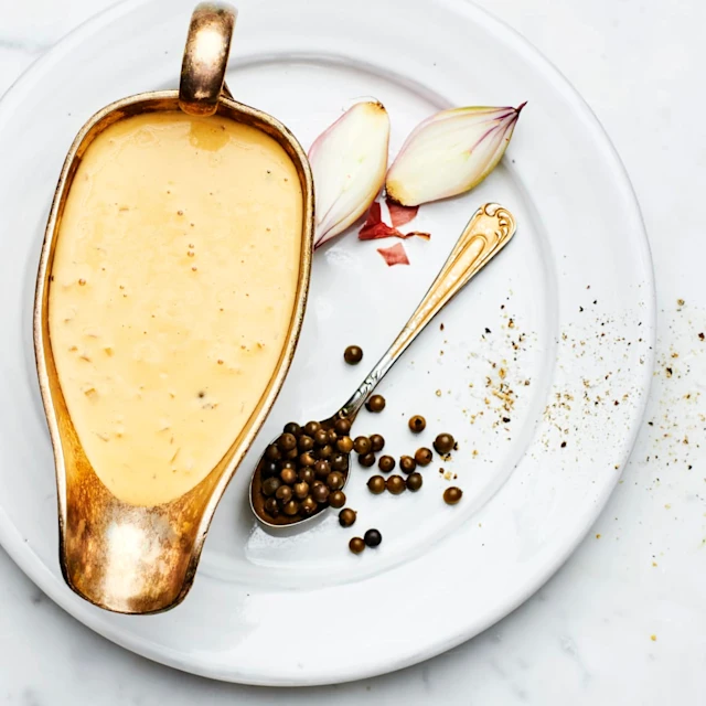 Grönpepparås i en skål som står på en vit tallrik. På tallriken ligger 2 stycken scarlottenlökar samt en sked med grönpeppar i. Det ligger 4 stycken scharlottenlökar med skal på till vänster om tallriken.