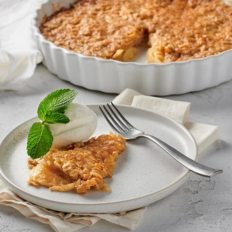En bit äppelpaj med gräddglass och mynta på en tallrik, med en pajform i bakgrunden.