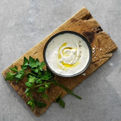 Fetaostsås i en liten svart skål som står på en träskärbräda. Olja är ringlad på såsen som en dekoration. Till vänster om skålen ligger en grön kvist, någon ört.