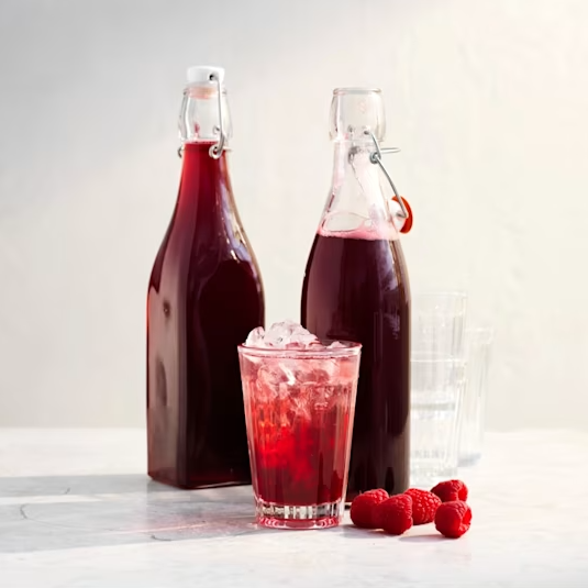 Två flaskor med röd dryck står bredvid ett glas med is och bär, på en ljus yta.