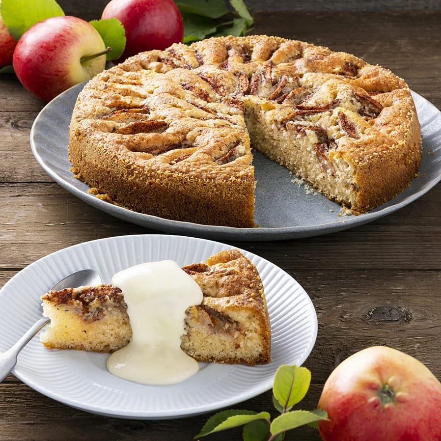 Äppelkaka på ett fat med en bit serverad på en tallrik, dekorerad med grädde och äpplen i bakgrunden.