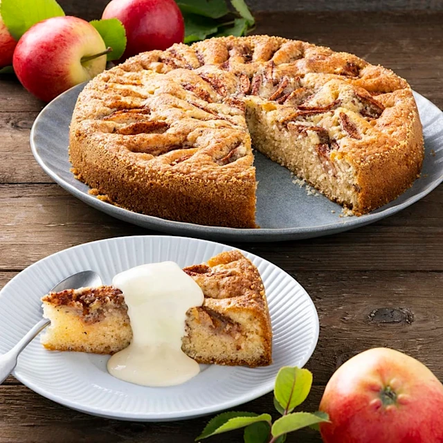 Äppelkaka på ett fat med en bit serverad på en tallrik, dekorerad med grädde och äpplen i bakgrunden.