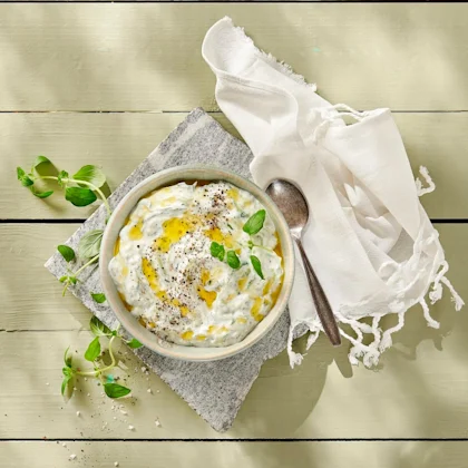 En skål med Tzatziki med olja och svartpeppar som dekoration. En vit tygservett ligger till höger om skålen, och en sked ligger på servetten.