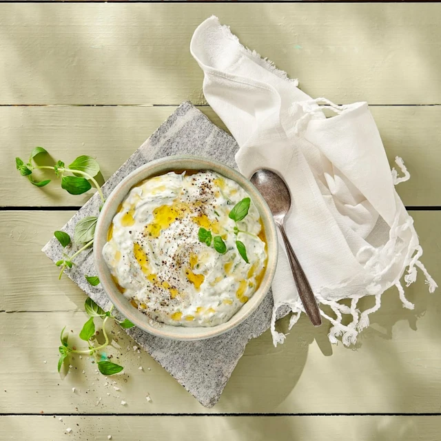 En skål med Tzatziki med olja och svartpeppar som dekoration. En vit tygservett ligger till höger om skålen, och en sked ligger på servetten.