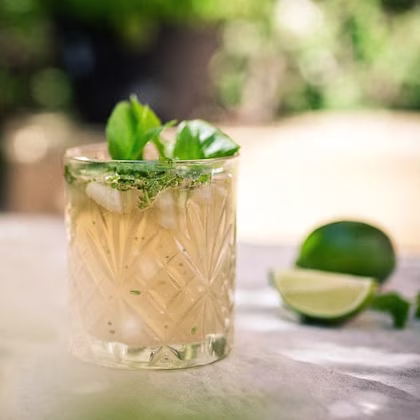 Kall drink med is och gröna örter i ett mönstrat glas, bredvid limehalvor på bord utomhus.