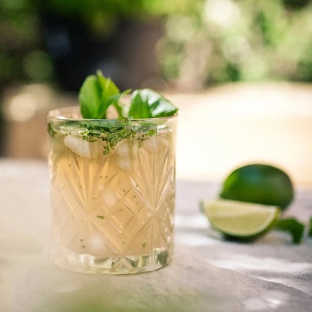 Kall drink med is och gröna örter i ett mönstrat glas, bredvid limehalvor på bord utomhus.