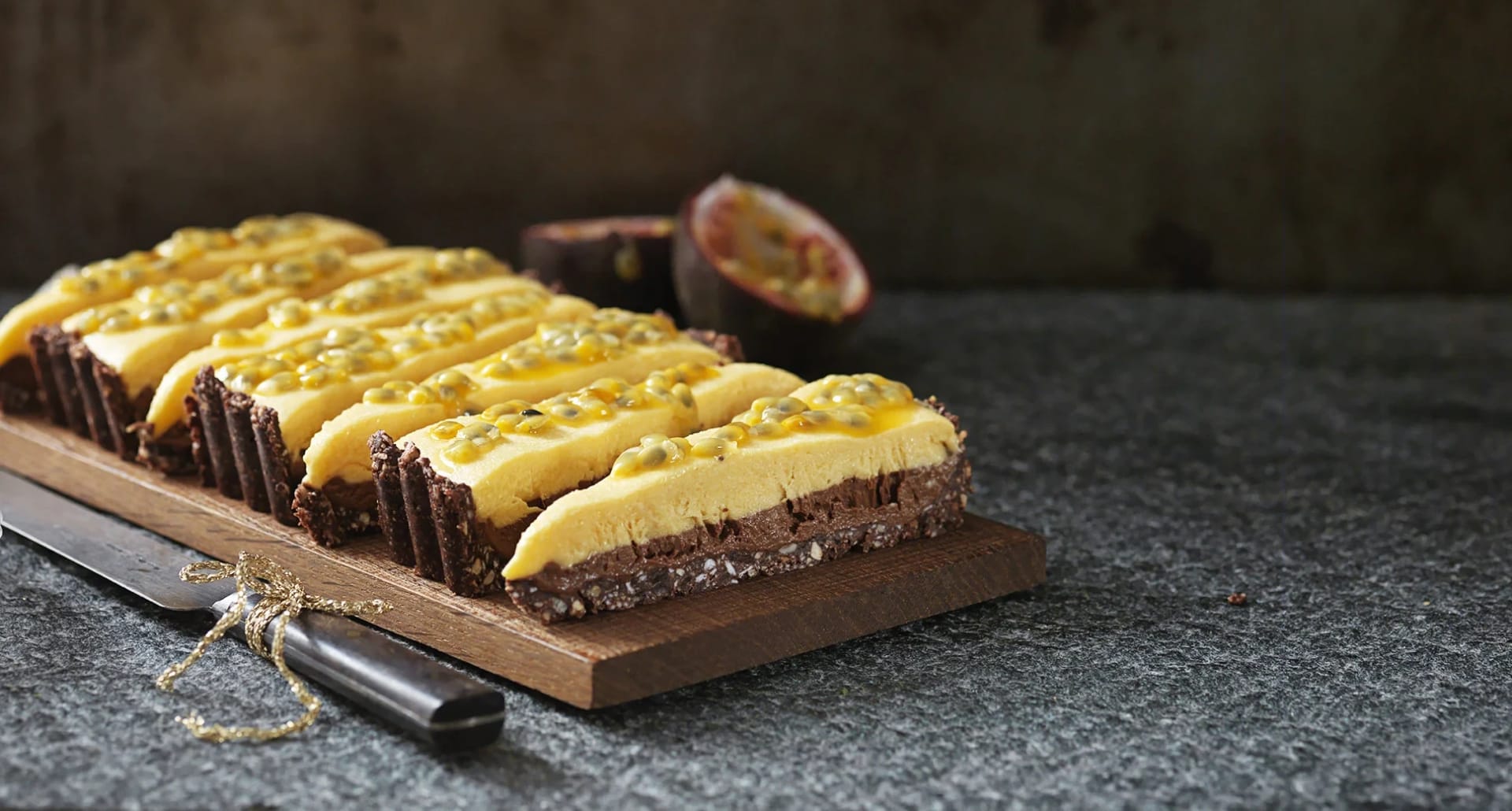 Fryst choklad- och passionstårta skuren i bitar på en skärbräda, toppad med passionsfrukt, med en kniv och en halverad passionsfrukt bredvid.