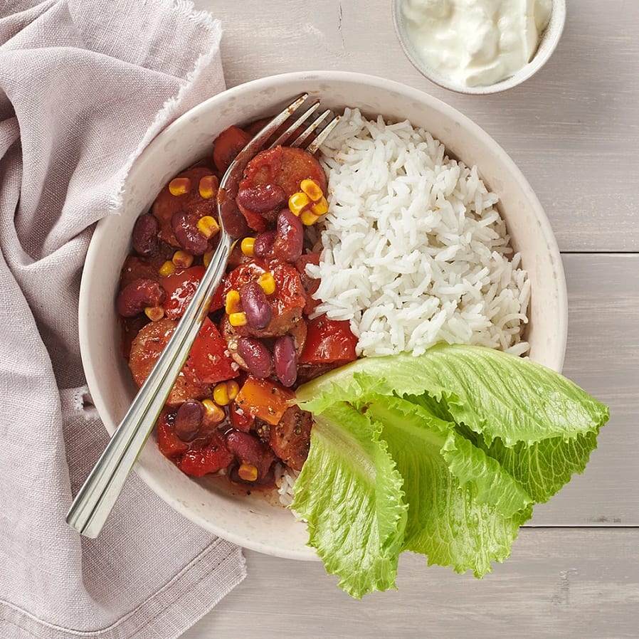 En vit djup skål med bitar av korv, majs och paprika. Serverad med ris och ett stort salladsblad. 