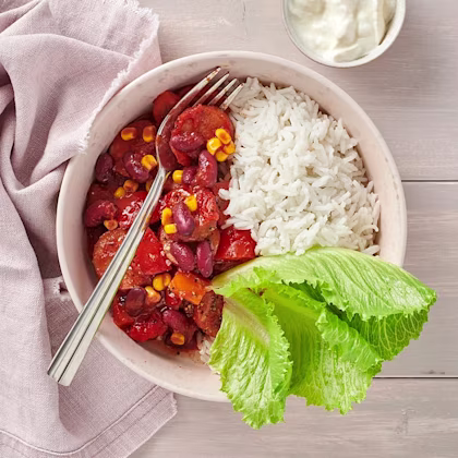 En vit djup skål med bitar av korv, majs och paprika. Serverad med ris och ett stort salladsblad.