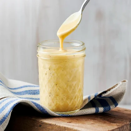 Gul mangorajasås i en transparent skål som står på en skärbräda i trä, samt en handduk som är beige med blåa ränder. Ovanför skålen är en sked med sås på som rinner ner i skålen.