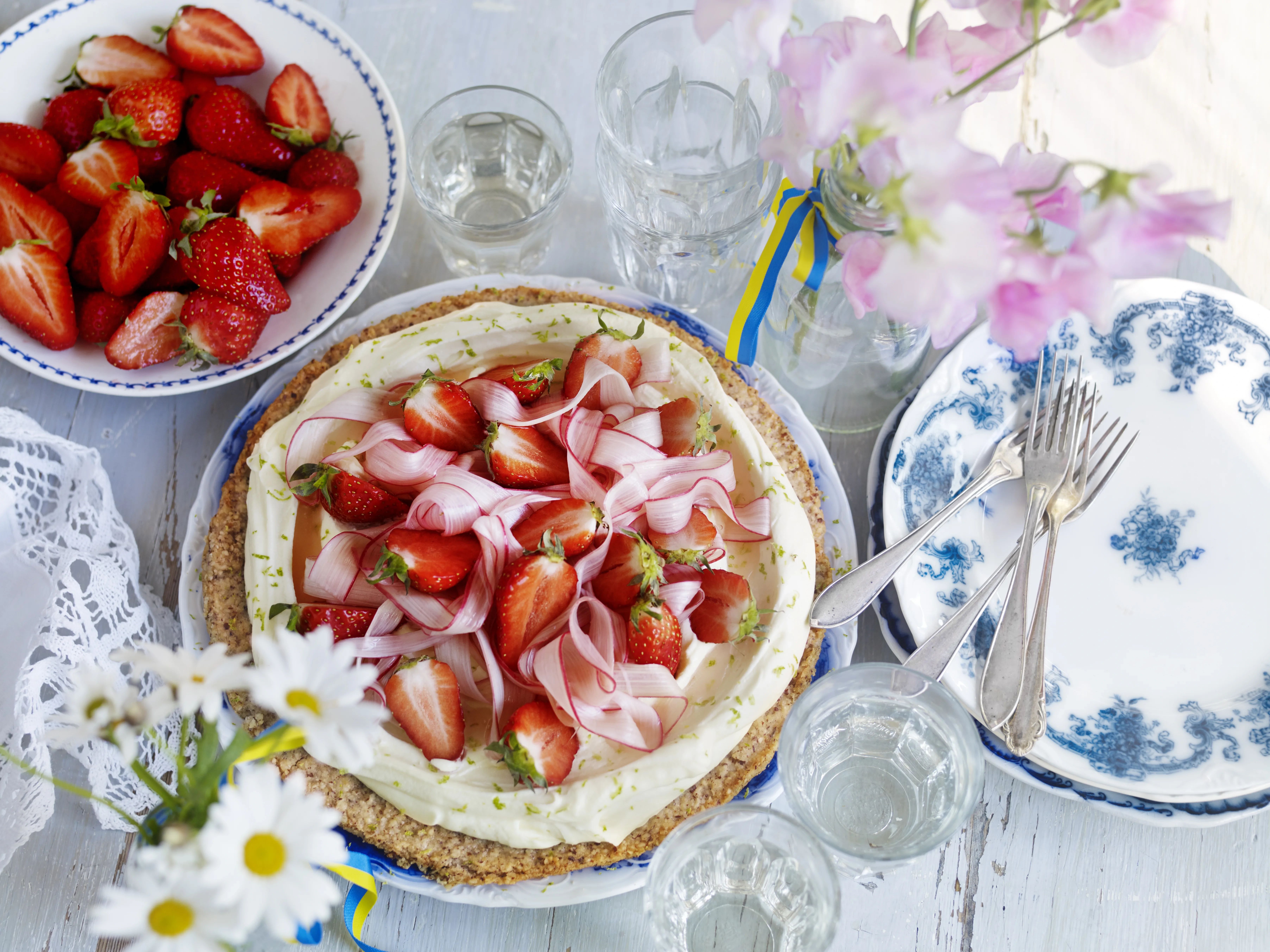 Sommartårta med grädde, jordgubbar och rabarber på bord med glas, tallrikar och blommor.