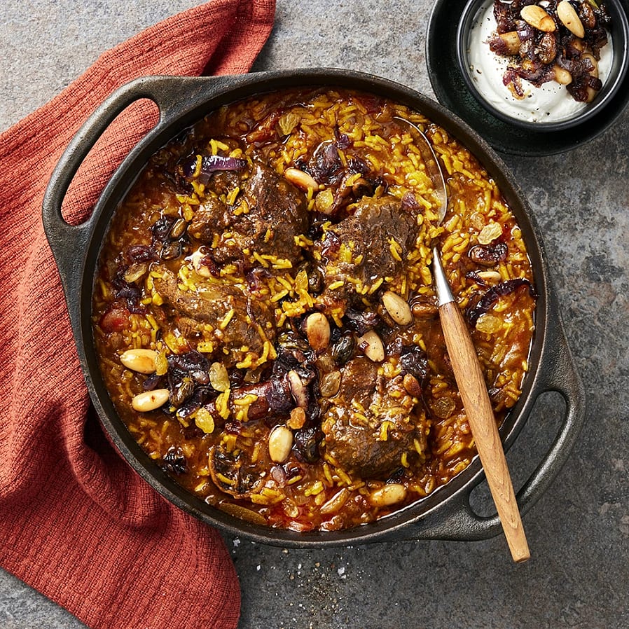 En gryta med ris, kött och nötter, serverad med en sked. En röd kökshandduk ligger bredvid.