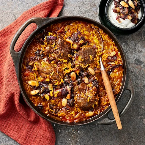 En gryta med ris, kött och nötter, serverad med en sked. En röd kökshandduk ligger bredvid.